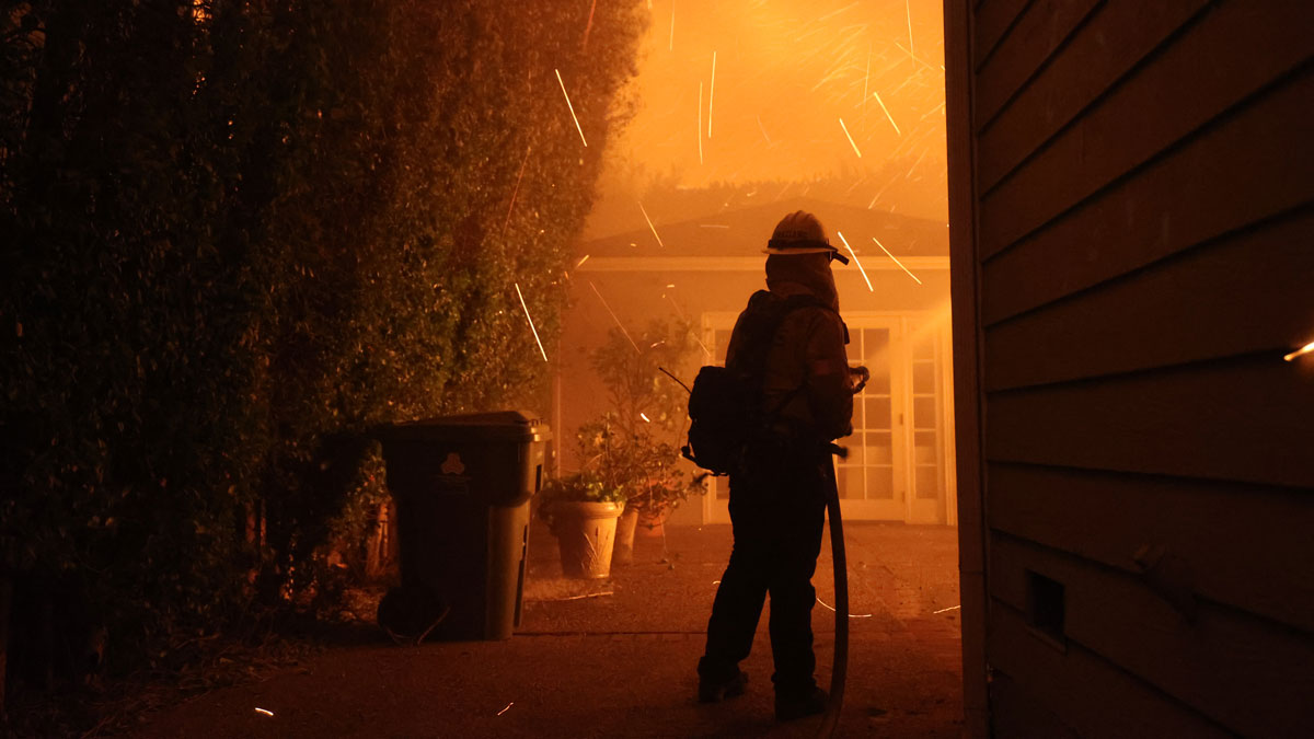 A firefighter, silhouetted against an orange fiery background and surrounded by flying sparks, sprays water at flames.