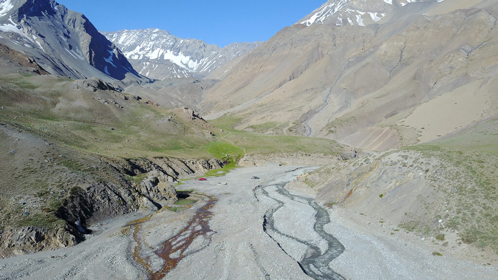 A rocky riverbed with two small streams surrounded by gray mountains. There are some green patches of plants and some snow on the peaks.