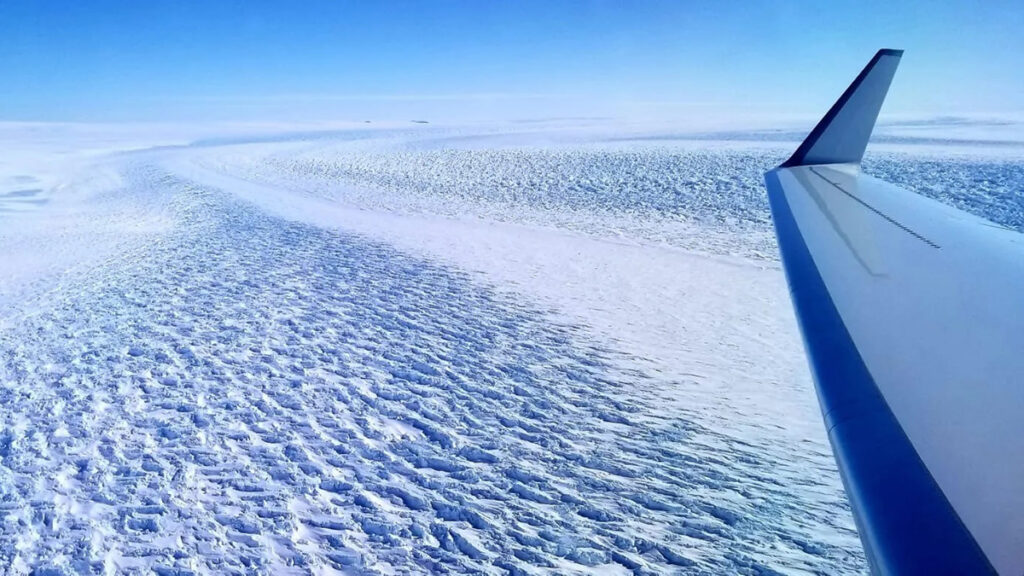 A glacier with ripples on top of it, as seen from a plane. A blue sky is visible.