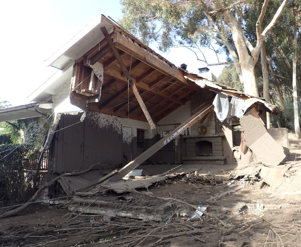 The inside of a half-demolished house that’s covered in mud and surrounded by downed tree limbs and debris