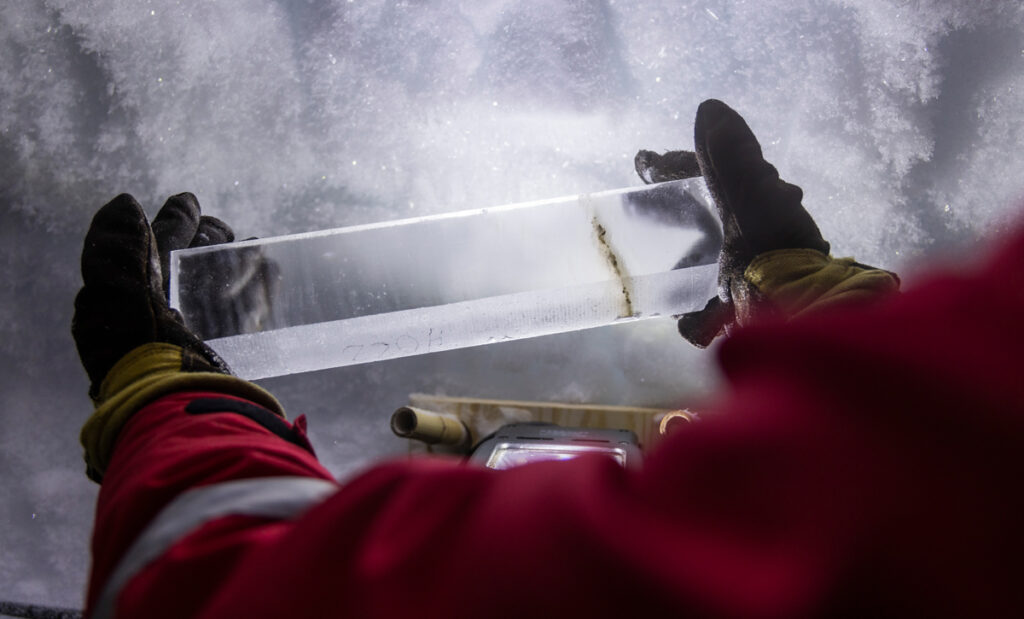 Two gloved hands hold a polished, translucent ice core that contains a clear, thin band of ash.