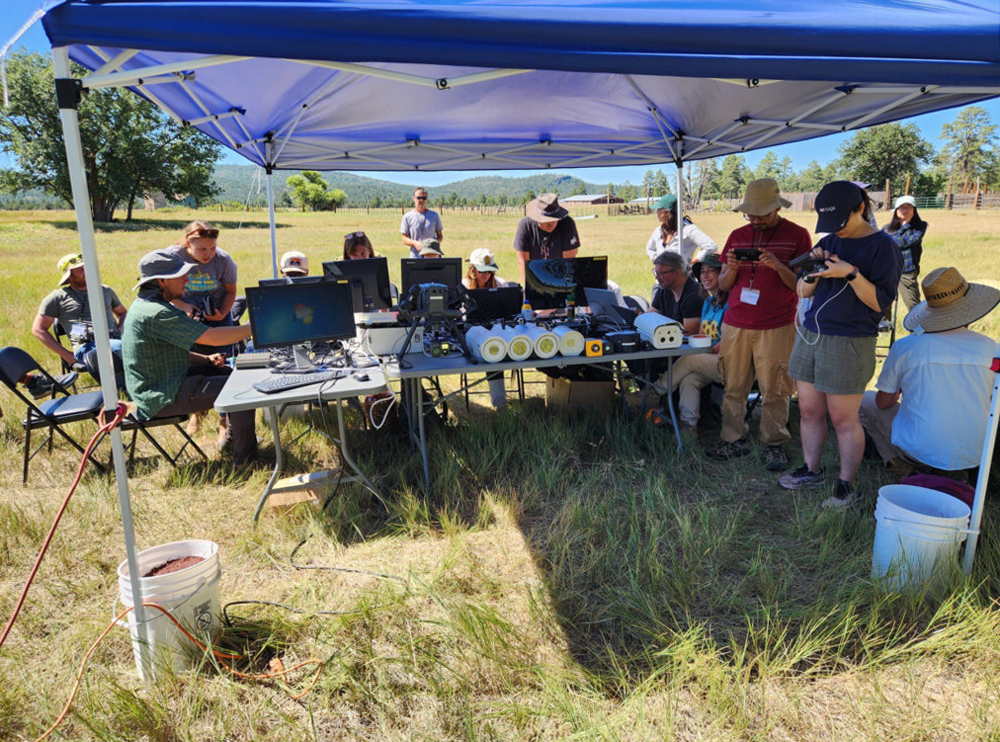 Participants in a workshop huddle around monitors and instrumentation under a large canopy set up in a sunny field.