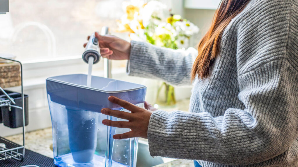 A woman in a sunny kitchen pours water from a kitchen tap into a filter pitcher.