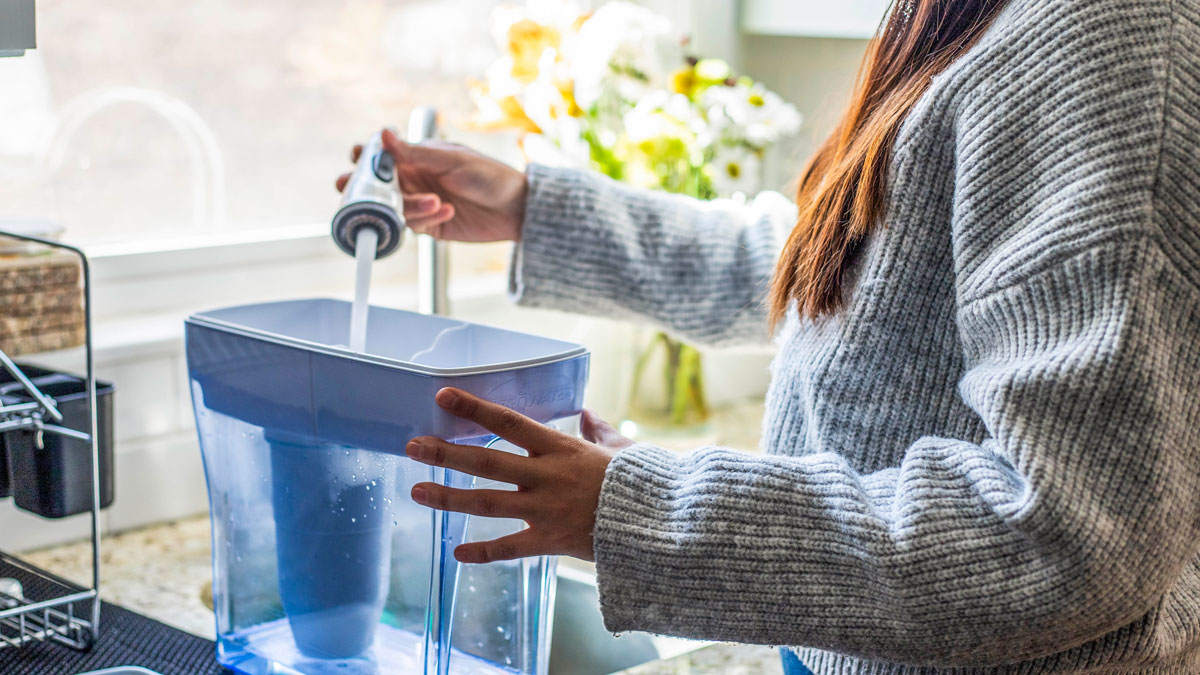 A woman in a sunny kitchen pours water from a kitchen tap into a filter pitcher.