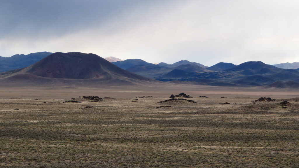 A mostly flat landscape is dotted with mounds, which give way to taller volcanic cones in the background.