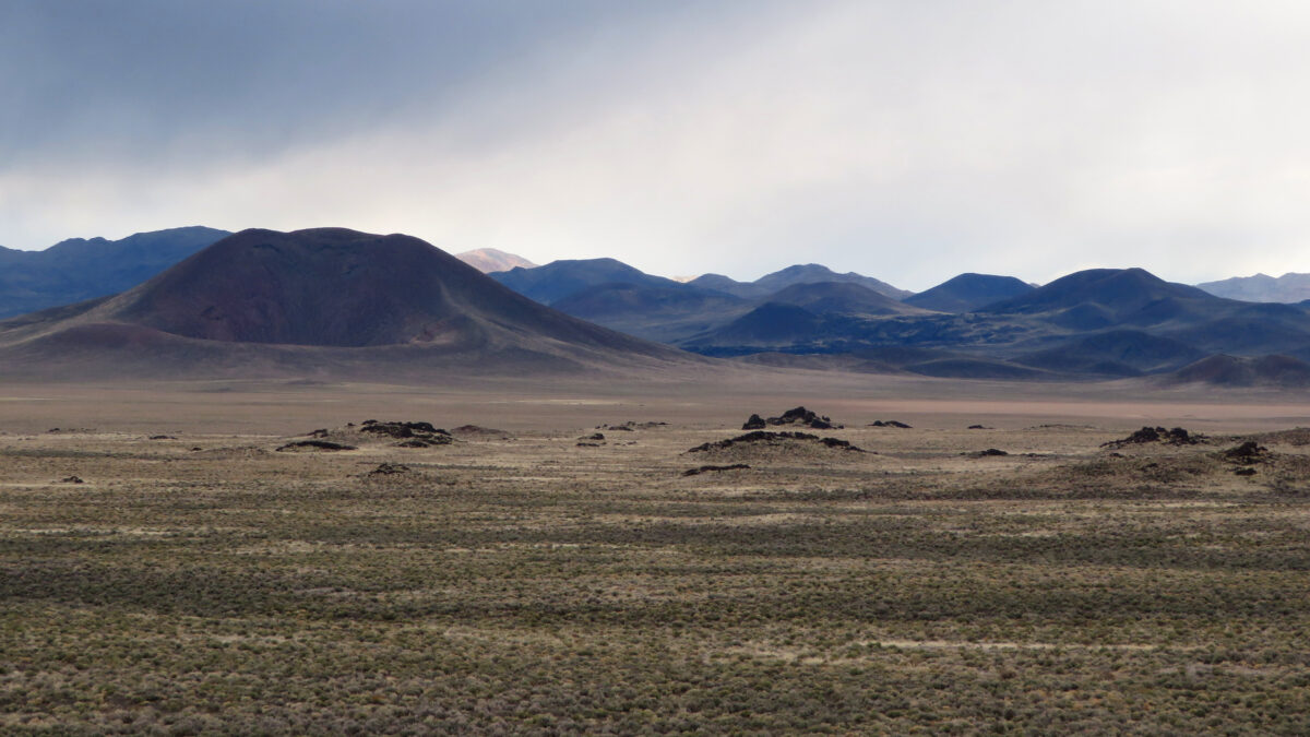 A mostly flat landscape is dotted with mounds, which give way to taller volcanic cones in the background.