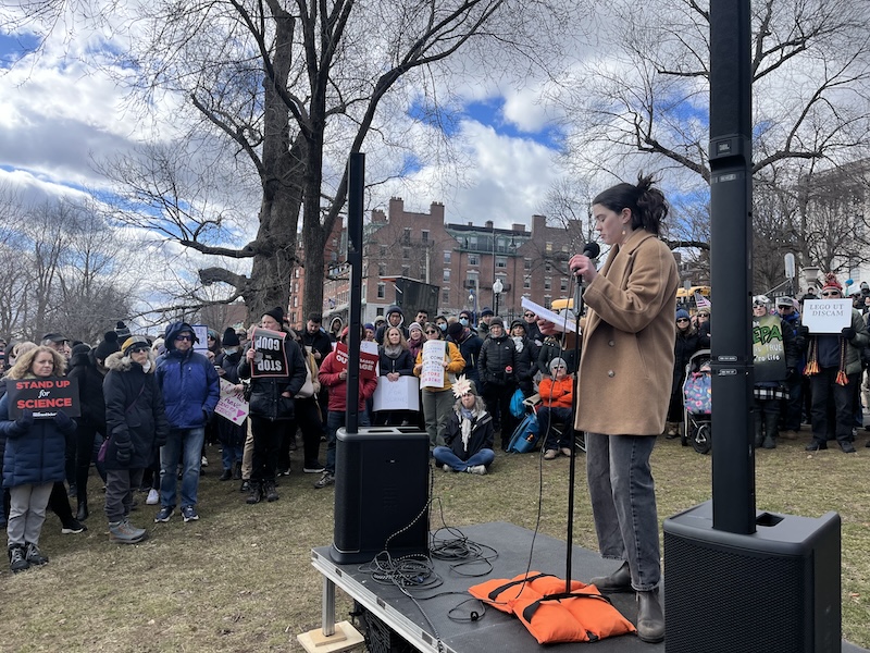 A person stands on a small stage speaking to a crowd.