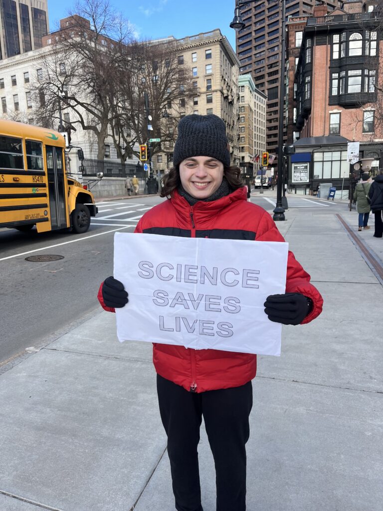 A person in a red coat and brown hat holds a sign that reads "Science Saves Lives."
