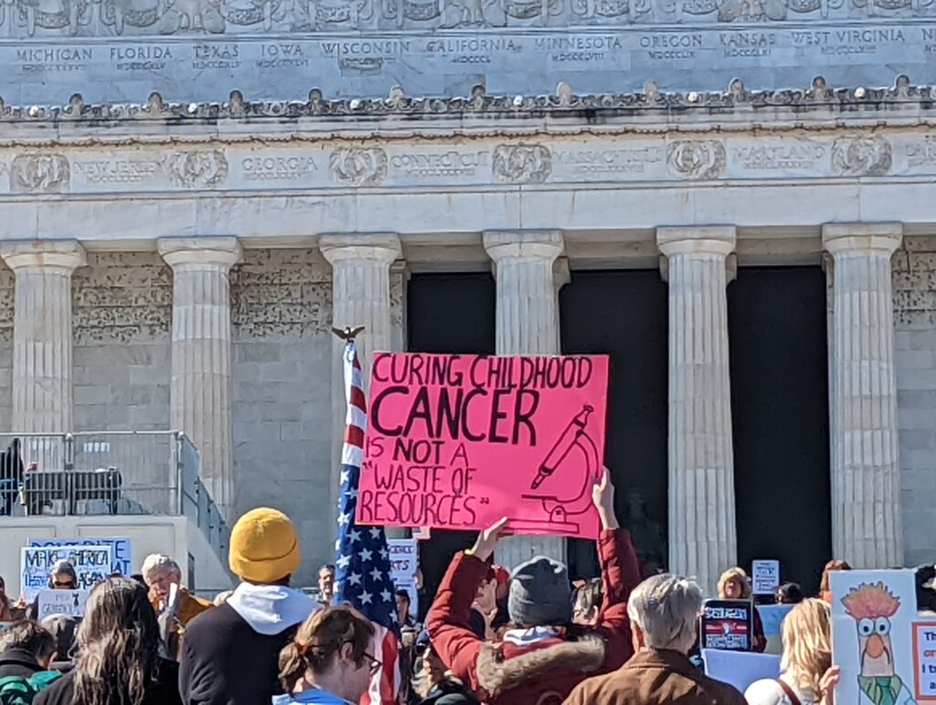 Protest in front of the Lincoln Memorial, with visible sign: Curing childhood cancer is not a "waste of resources."