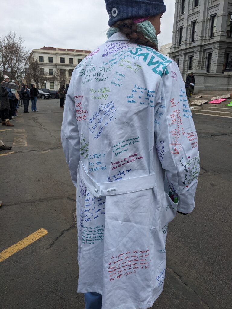 A woman wears a white lab coat. On the lab coat, others have written in colorful ink their reasons for choosing a science career.