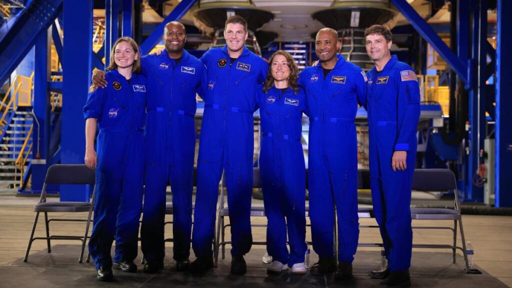 Six astronauts in blue flight jumpsuits stand in a line with arms around each other inside an industrial building.