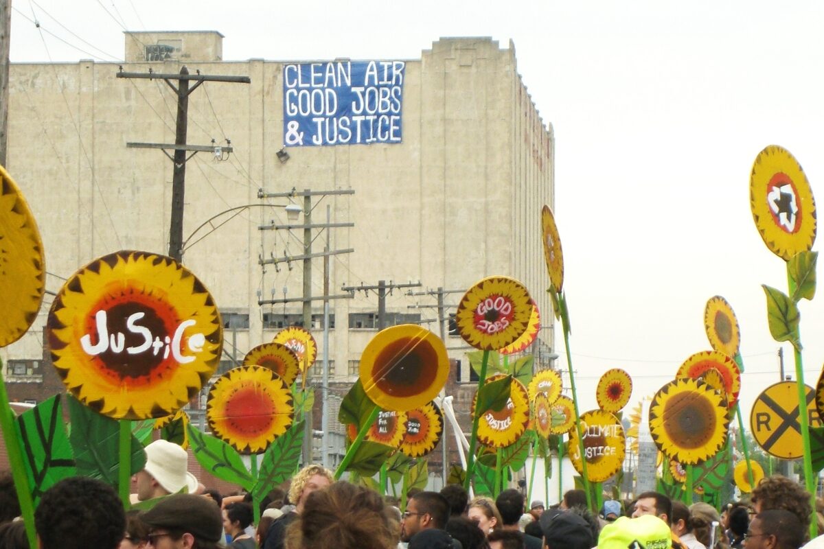 A group of people gathers with signs painted to look like sunflowers. On each sign is a word or phrase in support of environmental justice. A sign hanging from a nearby building says “Clean air, good jobs, & justice.”