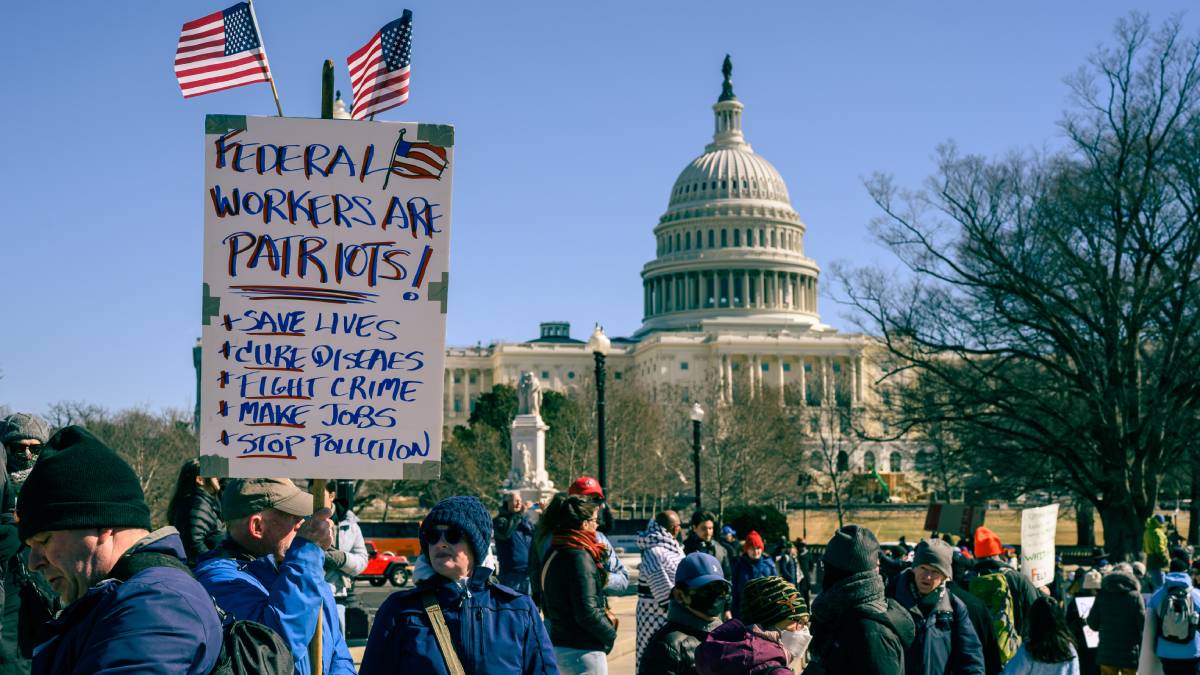 A person holds a sign reading, “Federal workers are patriots! Save Lives. Cure Diseases. Fight Crime. Make Jobs. Stop Pollution.” The person stands in a small crowd with the U.S. Capital Building in the background.