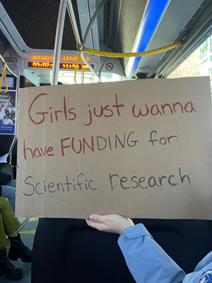 A handwritten sign reading “Girls just wanna have funding for scientific research.” In the background can be seen the interiori of a Metro bus.