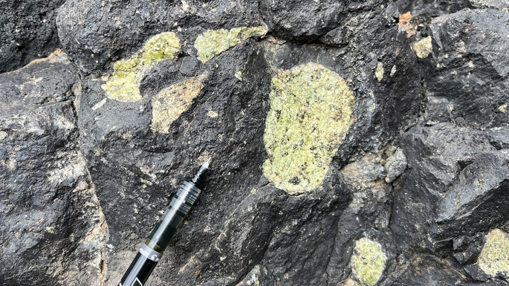 Close up view of the surface of dark-colored rock containing several large pale green crystals. The tip of a pen appears beside the rock for scale.