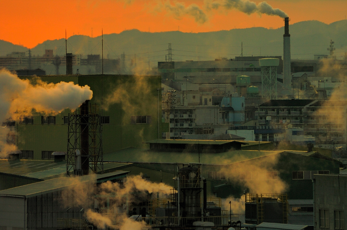 Several smokestacks at different levels pour smoke over a city. A mountain range and orange sky can just be seen peeking out over the tops of the buildings.