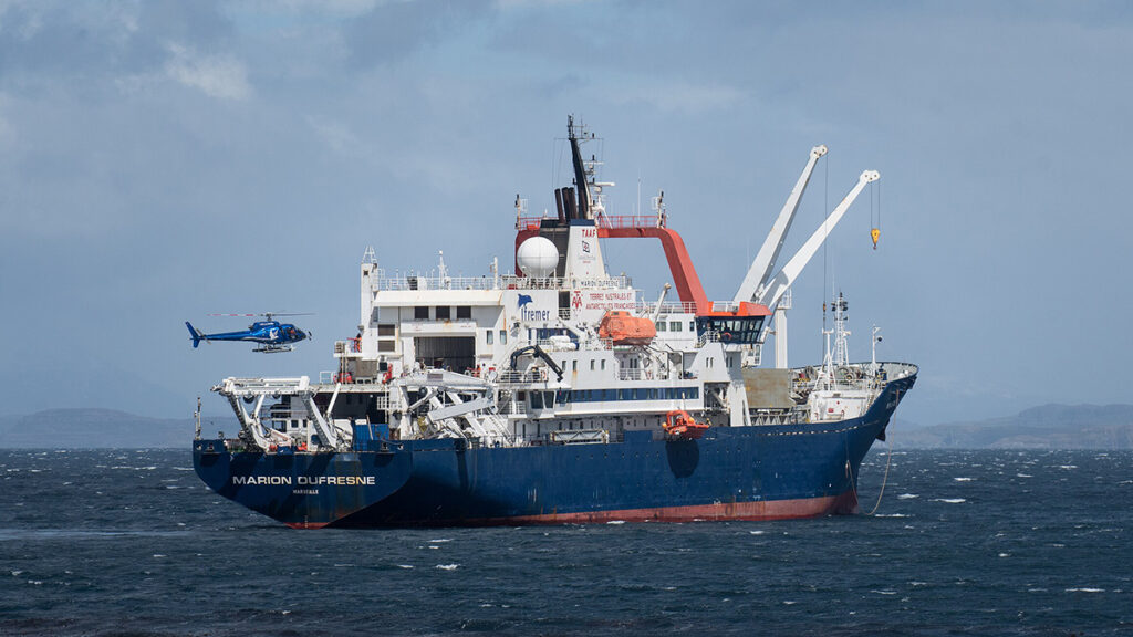 A large red, white, and blue ship with “Marion Dufresne” printed on the side is at sea, preparing for a helicopter to land on it.
