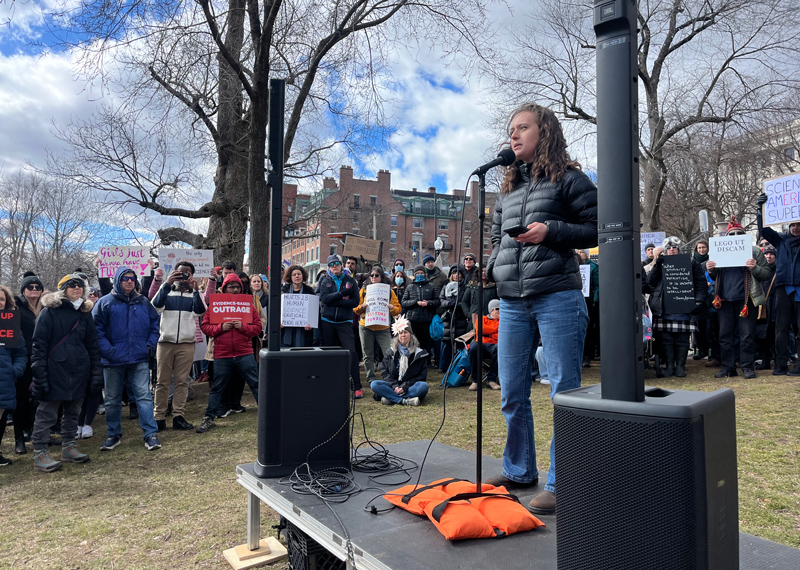 A young woman stands behind a microphone on a short platform upon which large speakers and cords also rest. In the background are many people, some of whom hold handwritten or printed signs.