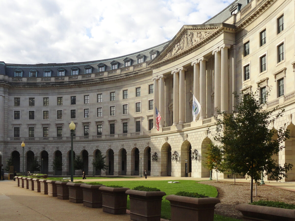 A curved building with classic pillars is seen from the sidewalk.