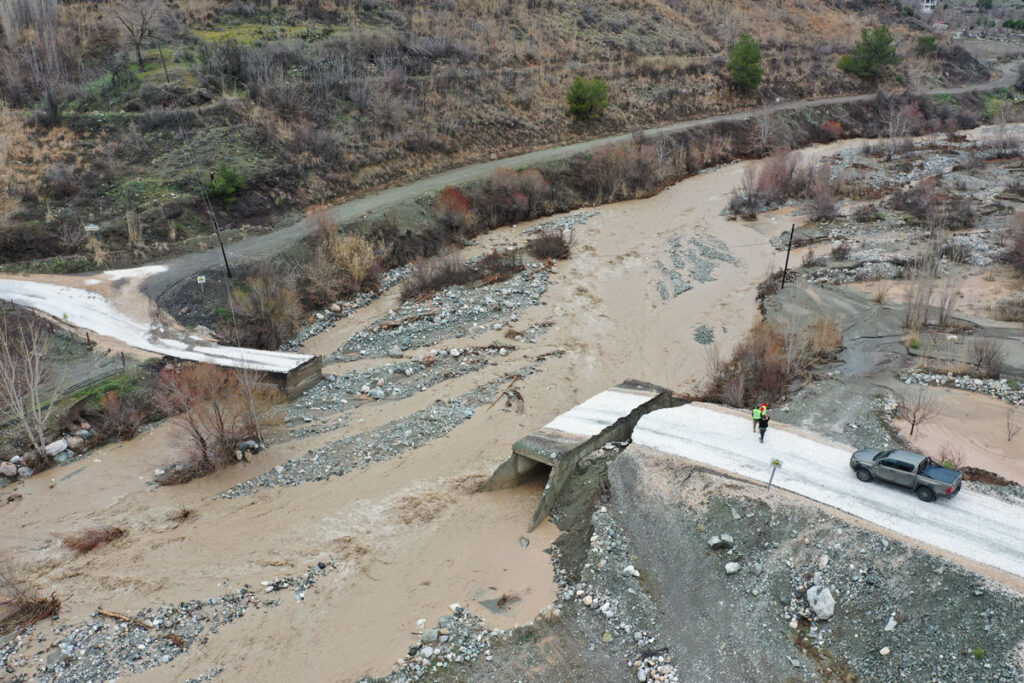An aerial view of a damaged bridge over a muddy river shows part of the bridge collapsed and washed away, with a pickup truck and an inspection worker on the remaining section.