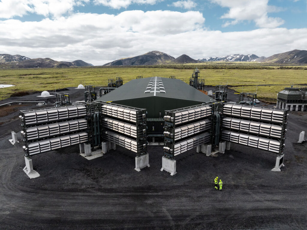 Wide view of an industrial-looking facility sitting atop a black ground surface in the foreground, with an open expanse of green vegetation and low mountains in the background.