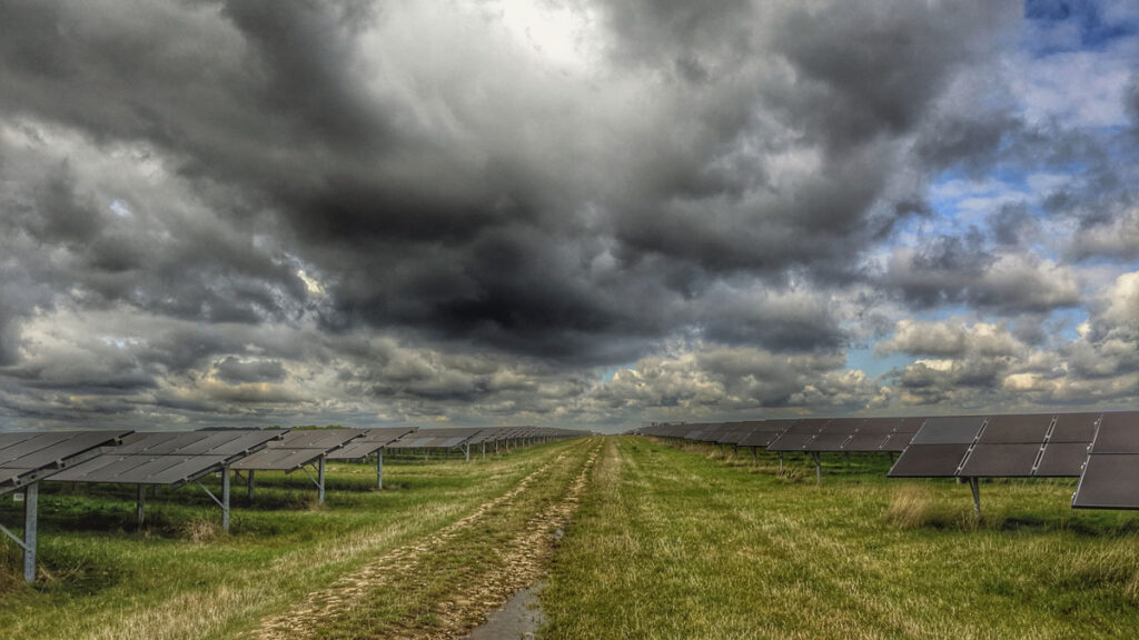 A stretch of green grass is visible between two rows of solar panels stretching into the distance. Gray clouds hang ominously over the scene.