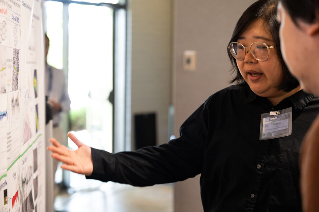 A standing scientist gestures to their research poster while explaining the results of their work to a colleague.