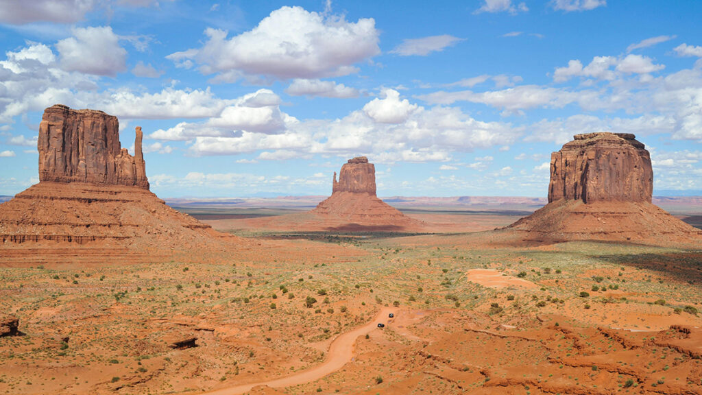 Three large rock formations tower over a desert. A blue sky and white clouds make up the background.