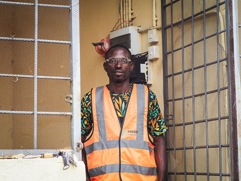 A man wearing glasses, a floral shirt, and an orange vest that reads “Prado Power” stands in the doorway of a building.