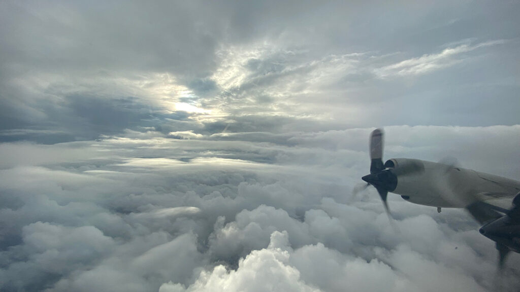 The wing of an airplane and a propeller are visible against a cloud-filled sky.