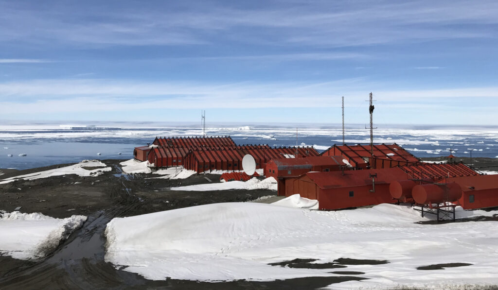 Several red buildings sit on an ice- and dirt-covered plateau overlooking an icy coastline.