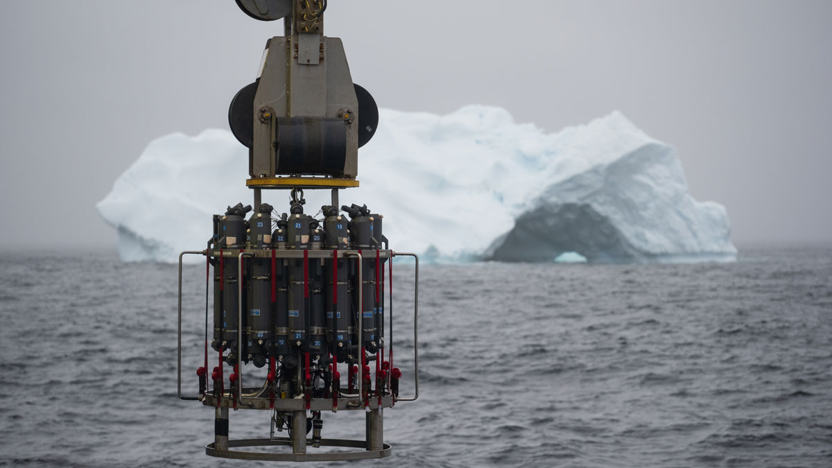 A metal tool consisting of many cylinders is being suspended over the side of a ship above gray ocean waters. An iceberg is in the background.