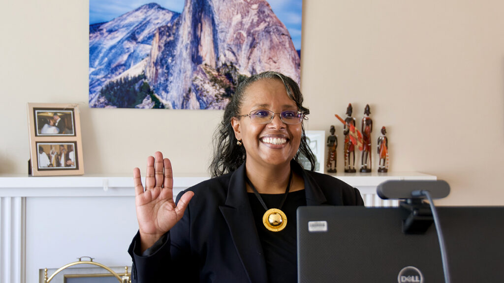 A woman stands in front of a computer monitor, smiling and holding up her right hand. She is wearing a large gold necklace, and behind her is a fireplace mantle with two framed family photos and a large photo of Half Dome.