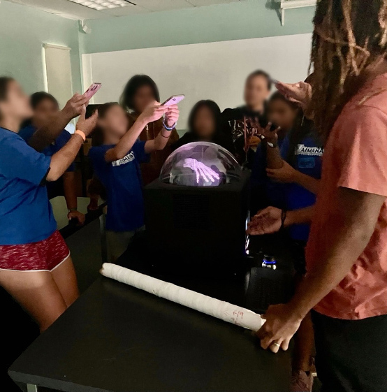 A group of middle school students surround a table with a coral core and a device showing a holographic image of a skeletal hand.