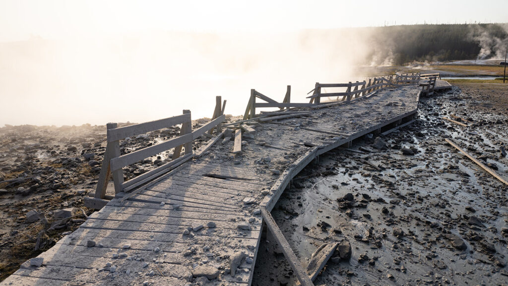 A heavily damaged section of boardwalk covered in rocks winds over muddy, debris-covered ground with steam rising from the ground in the background