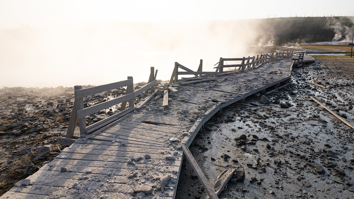 A heavily damaged section of boardwalk covered in rocks winds over muddy, debris-covered ground with steam rising from the ground in the background