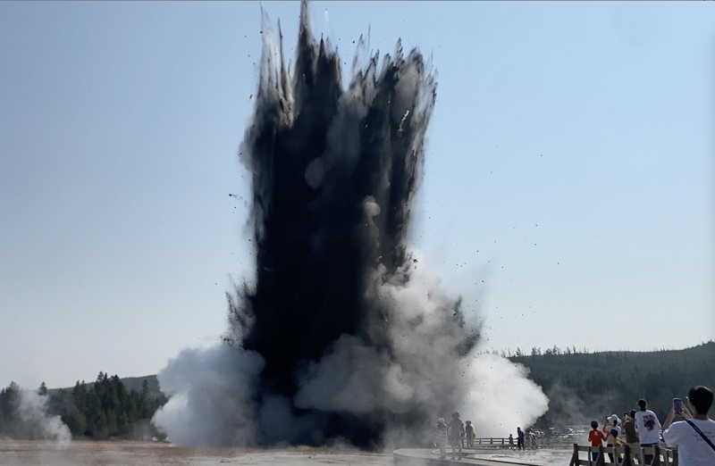 A tall plume of dark debris and white steam erupts from the ground as people in the foreground look on.