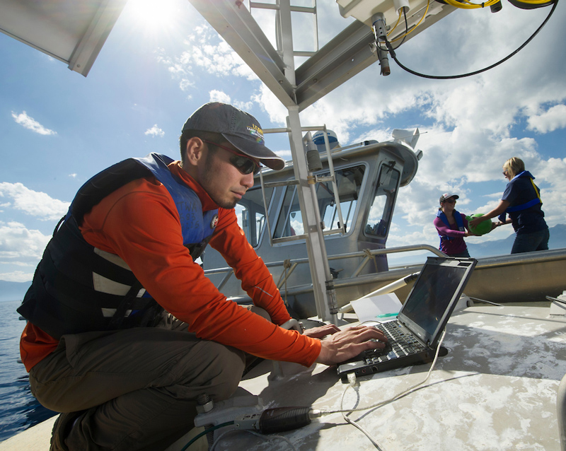 A scientist leans on a boat and works on a laptop with a sunny, blue sky and mountains in the background.