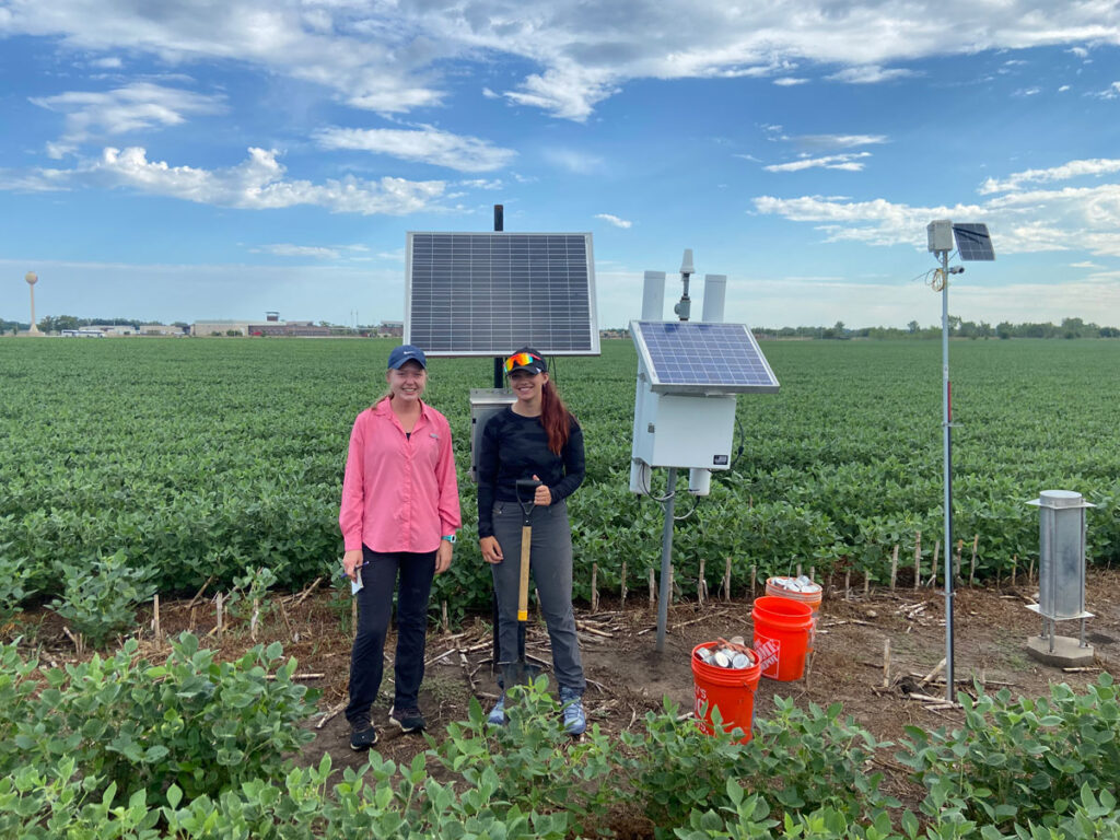 Two graduate students stand in an agricultural field beside scientific monitoring instrumentation topped with solar panels.