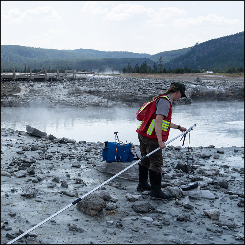 A National Park Service employee prepares water sampling equipment on the end of a long pole while standing near Black Diamond Pool.