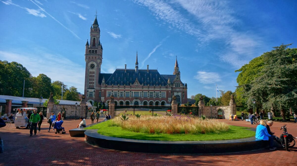 A large building with a tower is front-and center. People mill about outside the gate near some grass in the foreground.