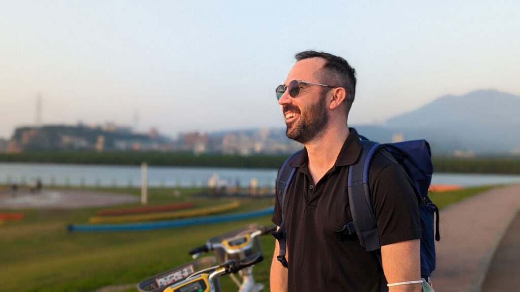 A man wearing sunglasses, a polo, and a backpack smiles while standing on a bike trail by the water.