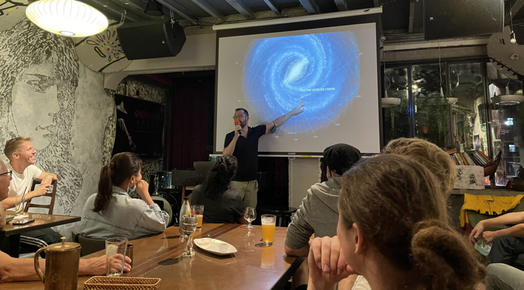 A man holds a microphone and points at a projection of a galaxy while presenting in a bar.