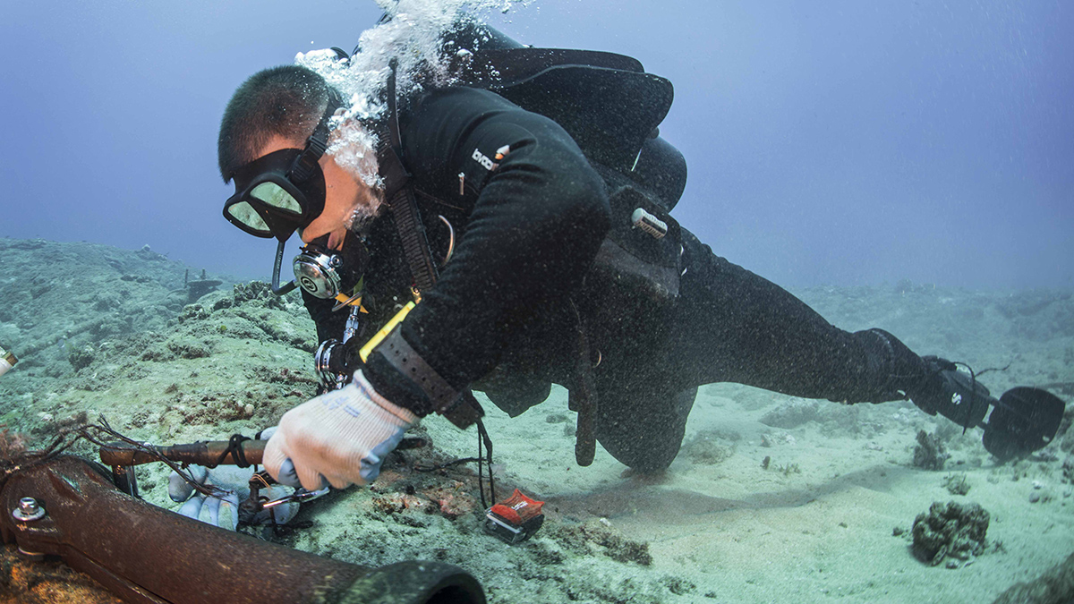 A diver, wearing goggles and an oxygen tank, uses a wrench to repair an underwater cable.