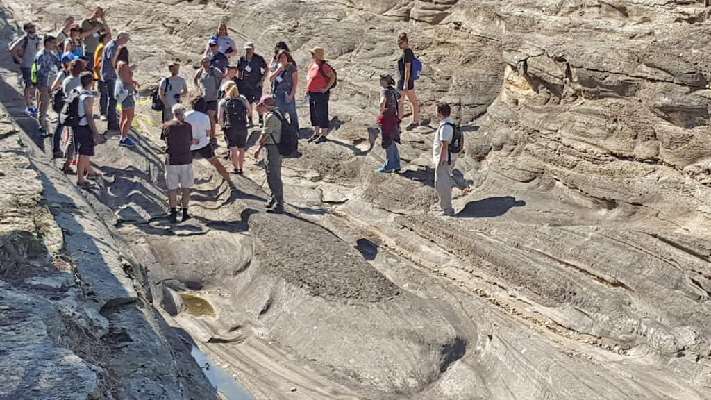 A group of people in the distance stand on a large gray rock outcrop with grooves carved by glaciers.