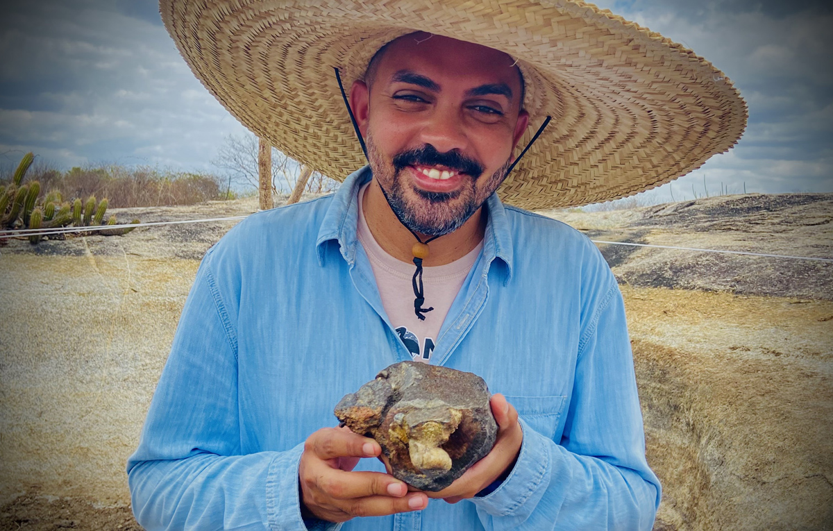 A person in a wide-brimmed hat smiles at the camera while holding a fossil.