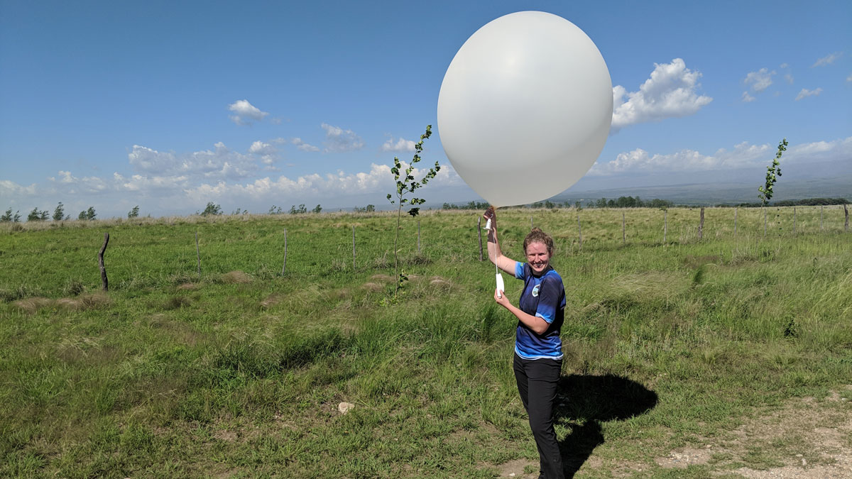 A woman wearing a blue T-shirt and black pants stands in a field of grass holding a large white balloon above her head.
