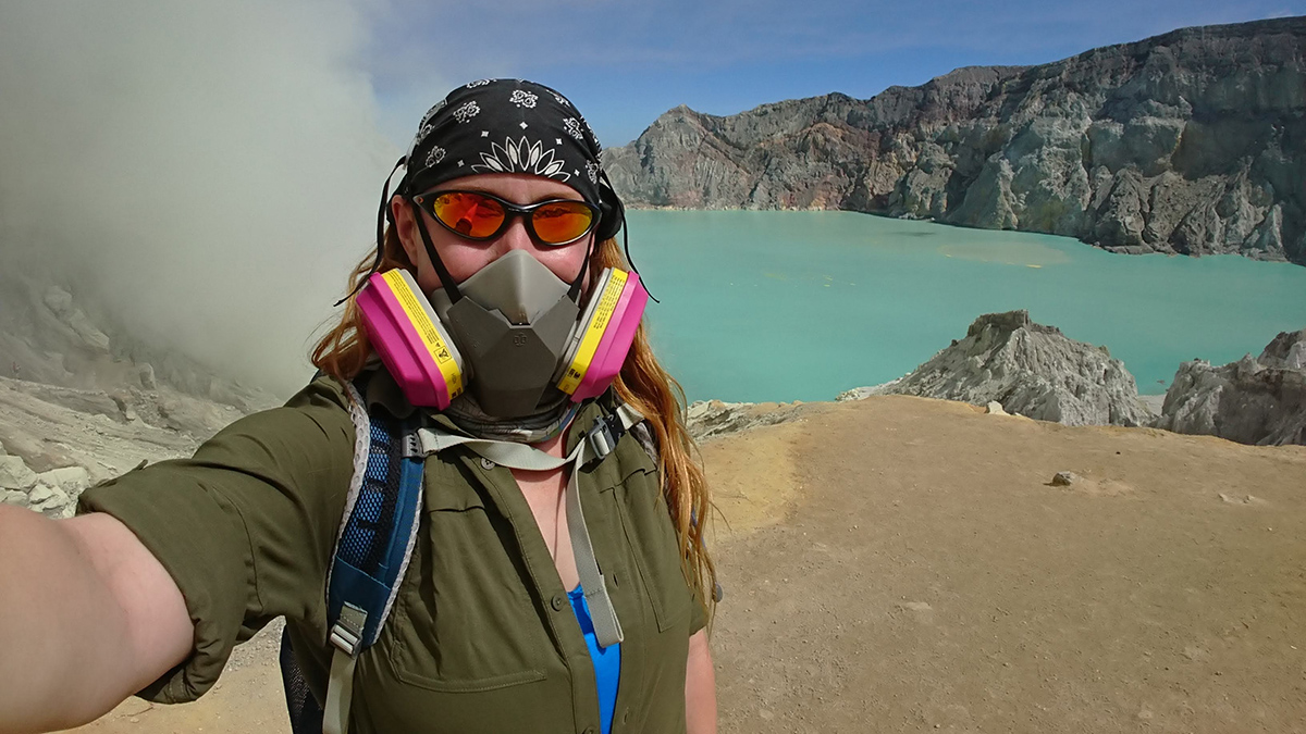 A person wearing a gas mask and sunglasses stands in front of a lake with steam covering the left side.