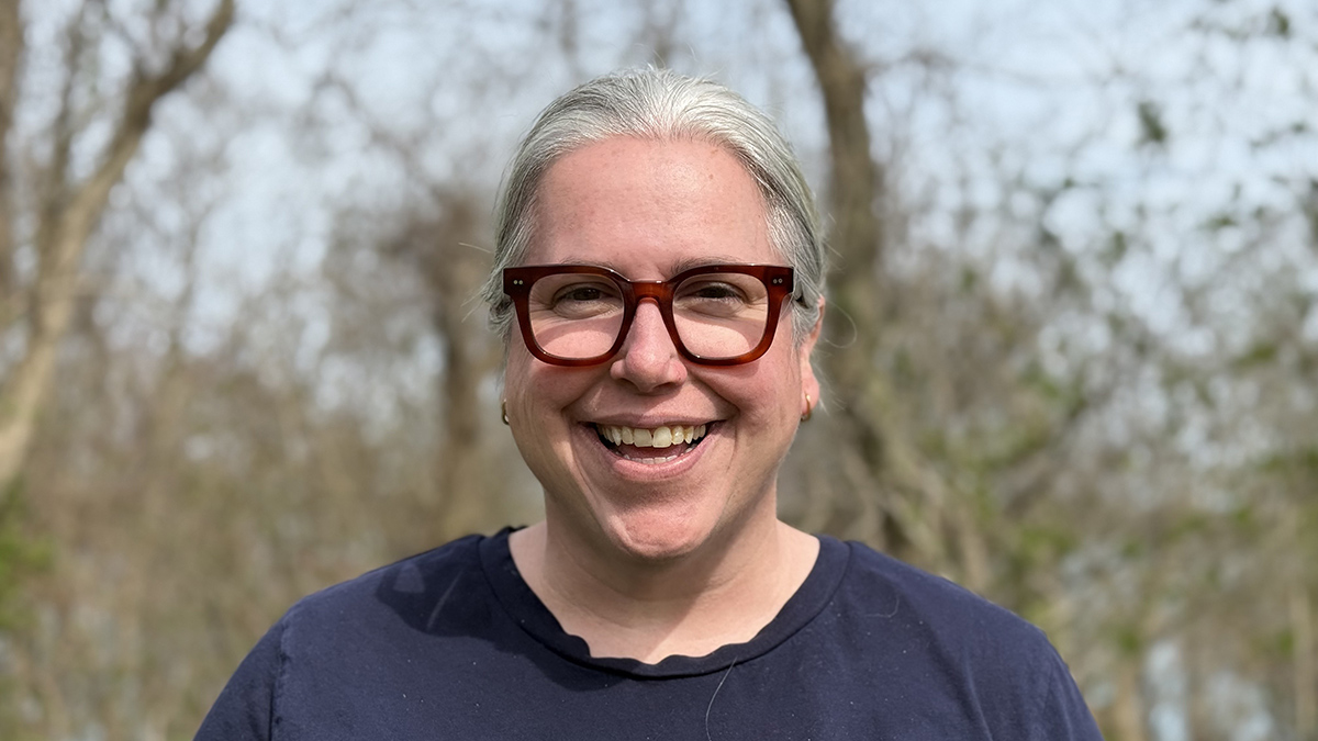 A woman wearing thick-rimmed glasses smiles at the camera. Trees are out of focus in background.