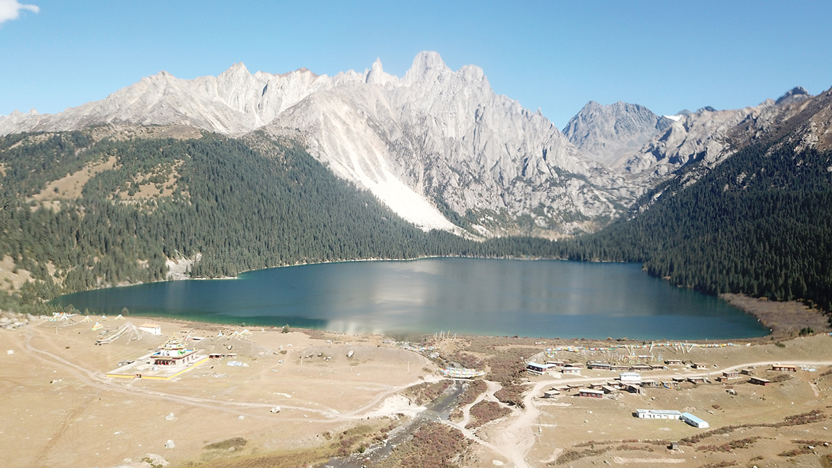 A clear blue lake lies between an expanse of dry dirt in the foreground and towering gray mountains whose lower reaches are covered in evergreen trees in the background.